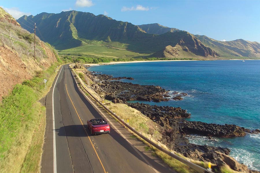 About Our Agency - Aerial View of a Red Convertible Driving on a Winding Road Along a Rocky Coast in Hawaii
