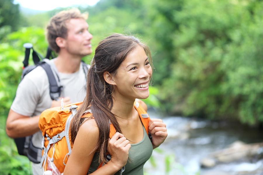 Contact Us - Young Couple Go Hiking Along a River Lined With Trees, Wearing Backpacks and Smiling
